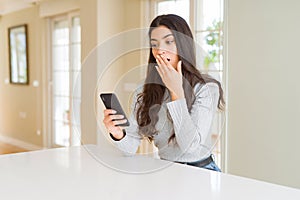 Young woman using smartphone sending a message cover mouth with hand shocked with shame for mistake, expression of fear, scared in