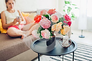 Young woman using smartphone sitting on couch at home. Living room decorated wih bouquet of roses