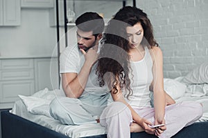 young woman using smartphone while upset man sitting behind on bed relationship