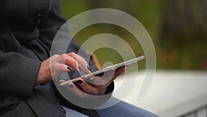 Young woman using tablet computer while sitting on bench in autumn park. Close-up of female hands touching of tablet