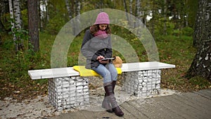 Young woman using tablet computer while sitting on bench in autumn park. Student with a Tablet Sitting on the Bench and