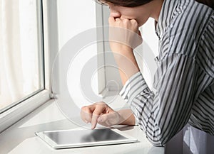 Young woman using tablet near window indoors, closeup. Loneliness concept