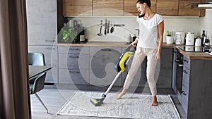 Young woman vacuuming carpet with cordless vacuum cleaner in the kitchen and dancing while listening to music