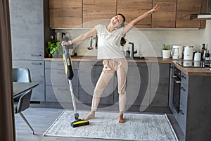 Young woman vacuuming carpet with cordless vacuum cleaner in kitchen and dancing while listening to music
