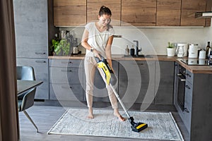 Young woman vacuuming carpet with wireless vacuum cleaner in kitchen. Household and useful technology concept.