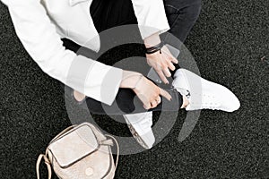 Young woman in a vintage jacket in black stylish ripped jeans with a gold trendy backpack in fashionable leather sneakers sits