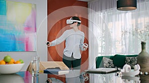 Young woman in a virtual reality helmet moving joysticks in her hands at home.