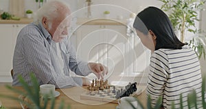 Young woman volunteer playing chess with senior man in kitchen at home bonding