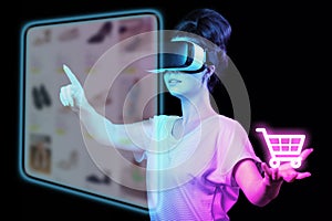 A young woman in VR glasses choice clothes at internet shop point at digital screen, holding neon virtual cart. Dark