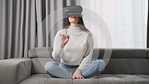 Young woman in VR glasses moves hand sitting on couch