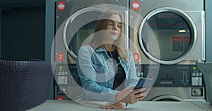 Young woman waiting for the clothes to be washed and listening to the music at the self-service laundry.