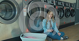 Young woman waiting for the clothes to be washed sitting on the floor and listening to the music at the self-service