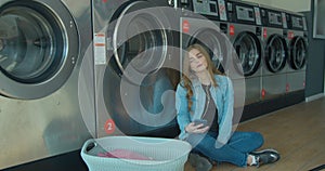 Young woman waiting for the clothes to be washed sitting on the floor and listening to the music at the self-service