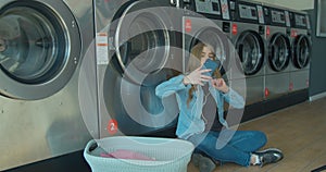 Young woman waiting for the clothes to be washed sitting on the floor and listening to the music at the self-service