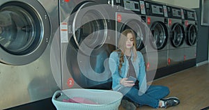 Young woman waiting for the clothes to be washed sitting on the floor and listening to the music at the self-service