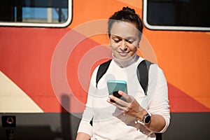 Young woman waiting train, using smart phone, checks mobile app, booking online tickets on internet. Railroad transport