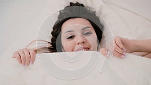 Young woman wakes up and have fun in bed. Girl start to hiding under sheets, making funny face and blowing hair