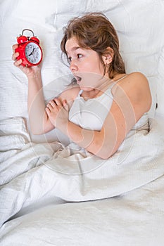 Young woman is waking up and looking at clock. She oversleep and is shocked photo