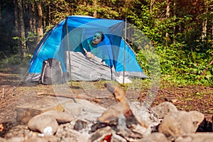 Young woman waking up unzipping tent by campfire in summer forest. Traveler enjoying recreational trip. Camping