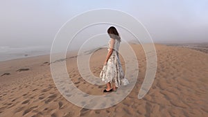 Young woman walking along sea shore sand beach in fog rainy day. Girl in long dress enjoy view on sand dune near ocean