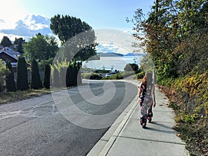 A young woman walking along the sidewalk towards the ocean in the distance. It is a beautiful neighbourhood on a pretty sunny day