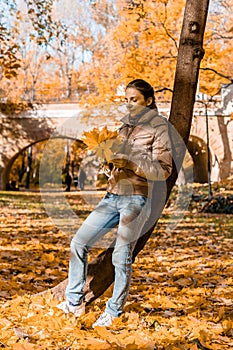 Young woman walking in autumn park. Pretty girl with orange leaves in hands leans on tree with autumnal foliage in background