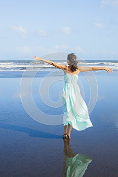 Young woman walking barefoot on empty beach. Full body portrait. Slim Caucasian woman wearing long dress. View from back. Water