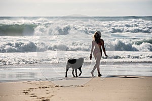 young woman walking barefoot on the sunny beach with her big dog