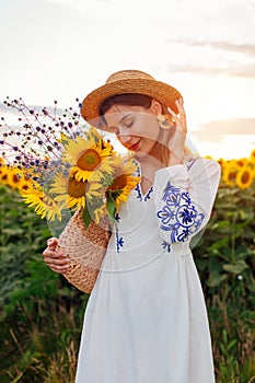 Young woman walking in blooming sunflower field at sunset smelling bouquet of flowers put in handbag