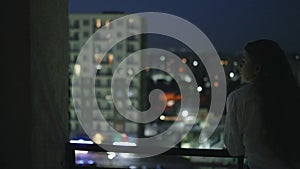 Young woman walking out from apartment enjoying night city view. Girl rests on balcony balustrade. Pretty lady looking