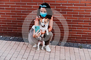 Young woman walking outdoors wearing protective mask, taking a picture with mobile phone of cute jack russell dog. New normal