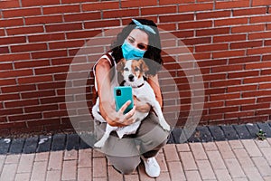 Young woman walking outdoors wearing protective mask, taking a picture with mobile phone of cute jack russell dog. New normal