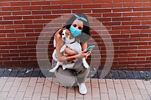 Young woman walking outdoors wearing protective mask, taking a picture with mobile phone of cute jack russell dog. New normal