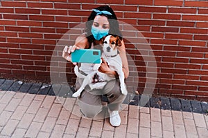 Young woman walking outdoors wearing protective mask, taking a picture with mobile phone of cute jack russell dog. New normal