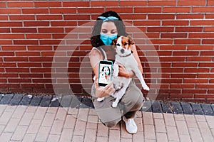 Young woman walking outdoors wearing protective mask, taking a picture with mobile phone of cute jack russell dog. New normal