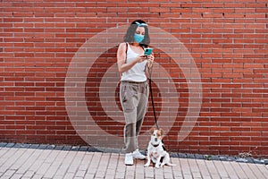 Young woman walking outdoors wearing protective mask, using mobile phone. cute jack russell dog besides. New normal concept