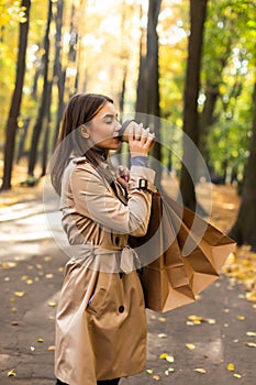 Young woman walking with shopping bag and drink hot coffee in autumn park