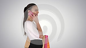 Young woman walking with shopping bags talking on mobile phone on gradient background.