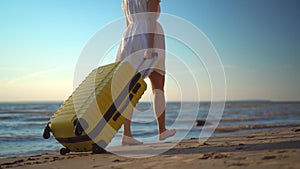 Young woman walks along the beach of the sea with a yellow suitcase closeup. A girl in a white dress walks barefoot on