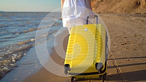 Young woman walks along the beach of the sea with a yellow suitcase closeup. A girl in a white dress walks barefoot on