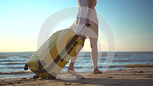 Young woman walks along the beach of the sea with a yellow suitcase closeup. A girl in a white dress walks barefoot on