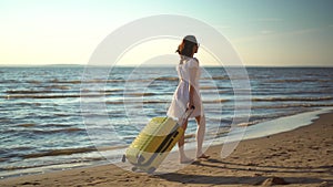 Young woman walks along the beach of the sea with a yellow suitcase. A girl in a white dress walks barefoot on the sand