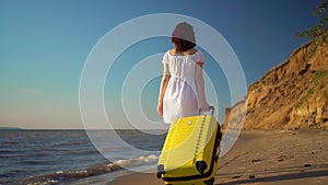 Young woman walks along the beach of the sea with a yellow suitcase. A girl in a white dress walks barefoot on the sand.