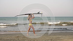 Young woman walks along the sea beach with a surfboard, slow motion