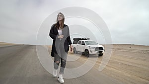 Young woman walks on highway, her car at roadside parked in desert. White SUV.