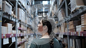 A young woman walks through the warehouse and looks for goods on the shelves. Rear view. Real time.