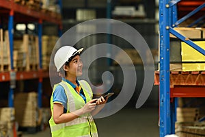Young woman warehouse worker in hardhats and reflective jackets using digital tablet and counting boxes on shelf
