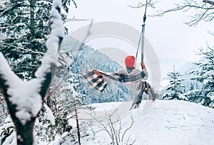 A happy young woman in warm clothes and Red Cap swinging on a forest tree swing with picturesque snowy mountain view. Wintertime