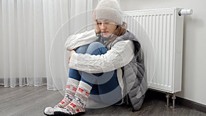 Young woman in warm clothes sitting at broken heating radiator in house. Concept of energy crisis, high bills, broken heating