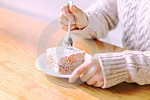 A young woman in a warm knitted sweater uses a teaspoon to cut a piece of delicious cake in a cafe on a brown wooden table. Cup of